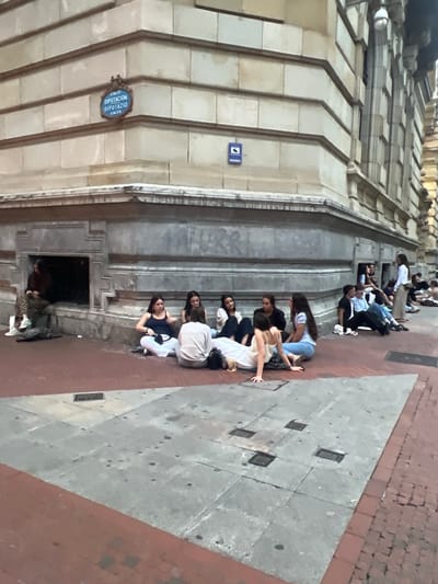 The image depicts a group of young people sitting on the ground outside a building with architectural detailing, likely in Bilbao, Spain, as suggested by a street sign visible on the building which reads "CALLE DIPUTACION DIPUTAZIO KALEA". The building has light-colored stone facade with horizontal bands and decorative elements. Several individuals are seated on the pavement in a semi-circular arrangement. One person is lying down on their side, propped up on their elbow. Another person is sitting in a recessed alcove of the building. In the background, more people are visible, some standing and others sitting. The ground is paved with a combination of red bricks and gray stone tiles. A few dark circular marks are visible on the gray tiles. There are also some metallic grates embedded in the pavement. The overall impression is a casual gathering of people in an urban environment.