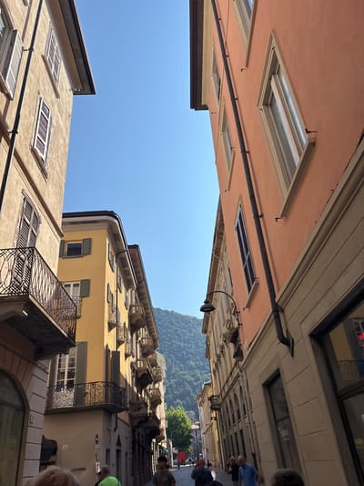 The image shows a street scene in Como, Italy, looking up at buildings against a clear blue sky. The architecture features a mix of pink, yellow, and beige facades with numerous windows, some with shutters, and ornate balconies. A dark pipe runs horizontally across the pink building. In the distance, a lush green hillside is visible. Several people are present, mostly blurred and seen from the back, suggesting they are walking or standing on the street. One person in the foreground appears to be wearing a green shirt. The sunlight suggests it is daytime, likely a sunny day. The perspective is from a low angle, looking upwards towards the sky and the upper floors of the buildings, creating a sense of looking up from within a narrow street. There is no visible text in the image.
