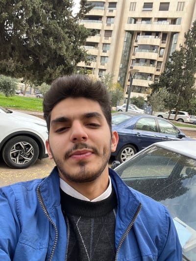 The image displays a headshot selfie of a young adult male, appearing to be in his late teens or early twenties, situated in the Kafr Sousa Municipality, Damascus, Syria. The male, positioned centrally in the foreground, has dark hair, dark eyes, and a short beard and mustache. He is wearing a white collared shirt beneath a black V-neck sweater with a diamond pattern, covered by a blue zippered jacket. He maintains direct eye contact with the camera and exhibits a slight smile with visible dimples on his cheeks.
Behind the individual, an urban street scene unfolds under what appears to be an overcast sky. To the left, a white modern SUV is partially visible, showing its front wheel and a section of its body. To the right, the rear section of a dark blue sedan is visible, with its driver's side window showing scattered debris. Further back, additional vehicles are parked along a street. The background features a prominent, tall, multi-story building characterized by a light-colored facade, numerous balconies, and expansive glass windows, indicative of modern architectural design. Green trees with dense foliage and shrubs are present throughout the background, bordering the street and the building. A modern streetlamp with multiple fixtures is visible between the parked cars and the background building.