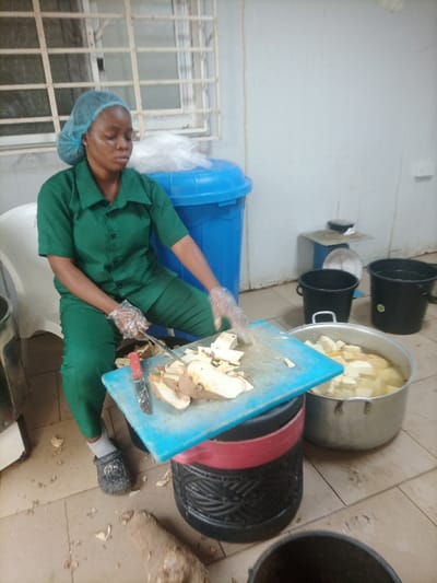 A woman wearing a green uniform and hairnet sits while cutting yams on a blue cutting board with a knife. She is wearing gloves. The yams have already been peeled and chopped. The cutting board is placed on a black and red stool. There is a pot of chopped yams in water next to her. In the background are buckets, a trash can, and a window. Yam peelings litter the floor.