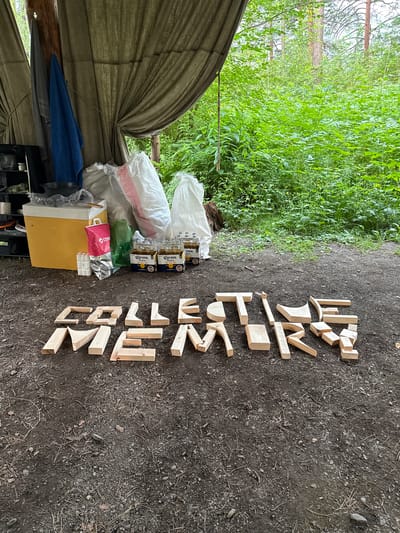 The image depicts an outdoor scene in a forest setting. In the foreground, wooden blocks are arranged on the ground to form the word "COLLECTIVE MEMORY". Behind the blocks, to the left, is a yellow cooler, various bags, a red bag, and several bottles of Corona Extra beer. Further in the background, tents or tarps are visible, with lush green foliage and trees surrounding the area. The ground is covered in dirt and small stones. There are no people or animals visible in the scene. The specific location in Cheposh, Russia cannot be determined from the visual information provided.