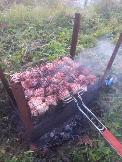 In the city of Polevye Ineli, Russia, a close-up, eye-level shot captures meat grilling over coals. The grill, made of rusted metal, is filled with numerous pieces of raw, pink meat arranged on a wire rack. The meat appears to be chicken or pork. The grill is placed outdoors, surrounded by green grass and other vegetation. Smoke rises from the grilling meat, indicating it is in progress. A blue can is visible on the ground to the right of the grill. The scene suggests a casual outdoor cooking event.