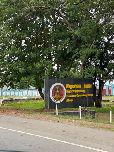The image shows a sign for the "Nigerian Army Peacekeeping Forward Operating Base Abuja". The sign is black with yellow lettering and a circular emblem featuring a lion and a star. It is situated outdoors, with trees and greenery surrounding it. In the background, there is a blue and white fence and some red structures. The scene appears to be a road or pathway leading up to the base. The overall environment suggests a military installation within a somewhat rural or suburban setting.