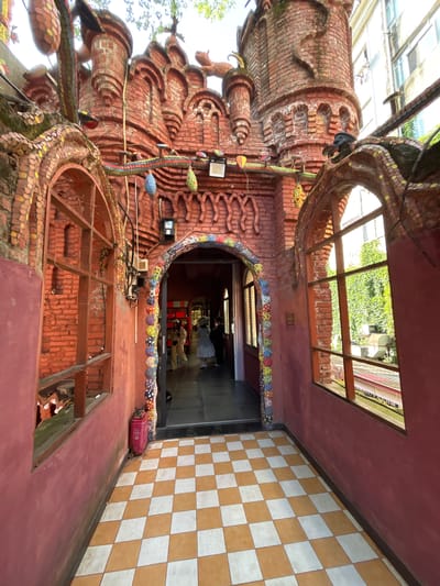The image captures the exterior of a building in Baqiao, China, characterized by unconventional brickwork and mosaic decorations. A large, ornate brick structure with decorative elements resembling scales or coils dominates the left side of the frame, extending upwards. Two window openings, one at the top and one in the lower section, show an open frame structure. The area to the right of the brick structure features a checkered tile pattern on the wall and a dark opening that appears to be an entryway or passage. Inside the opening, people and objects, including what looks like red furniture, are partially visible. A small, reddish object sits near the base of the checkered wall. The overall impression is of a unique architectural style with a mixture of brickwork, mosaic art, and modern elements.