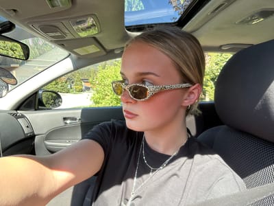 A woman with light brown hair and sunglasses is sitting in the driver's seat of a car. The car is parked outside and there are trees visible through the windshield. The woman is wearing a black shirt and a silver necklace. The interior of the car is gray. The location cannot be determined to be Whistler, Canada from the image provided.