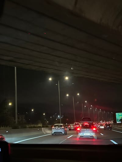 The image captures a night scene on a multi-lane highway, viewed from inside a vehicle. The perspective is rotated, showing a dark, textured wall or barrier on the left side, running vertically along the frame.
On the right side of the image, a busy highway stretches into the distance, filled with vehicles. Numerous red taillights from cars in traffic are visible, indicating either slow-moving or stopped conditions. Bright streetlights line the highway, casting illumination on the road and surrounding area.
In the foreground, a light-colored car (possibly white or silver) is prominent, with its rear facing the viewer. It appears to be equipped with a roof rack. Another car is visible directly behind it, and a long line of additional vehicles extends further down the road.
A green highway sign, partially obscured and indistinct, can be seen in the upper right portion of the image, suggesting an upcoming exit or destination. The dark sky confirms it is nighttime. The weather appears clear, with no visible precipitation or fog. No people are distinctly visible, and any text on the distant highway sign or vehicle license plates is illegible due to distance and image quality. The overall impression is one of night-time traffic on a major road.