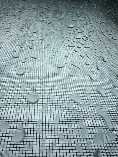 The image shows a close-up, high-angle view of water droplets on a black mesh surface, possibly a window screen. The mesh is composed of small, uniform squares. The water droplets are scattered across the surface, varying in size and shape. Some droplets are small and round, while others are larger and more elongated, some appearing to be running down the mesh. The lighting creates shadows cast by the water droplets onto the mesh, highlighting their three-dimensional form. The background is out of focus, appearing as a lighter, textured surface, suggesting this mesh is in front of something. The scene is static and devoid of human presence. The location is identified as Khazar Raion, Azerbaijan.