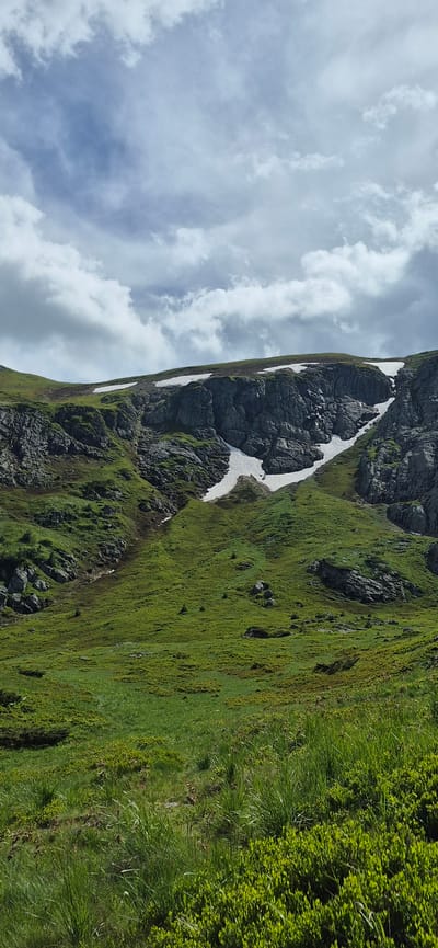 The image depicts a mountain landscape with green grassy slopes, rocky outcrops, and patches of snow. The sky above is partially cloudy. The terrain appears rugged and natural, characteristic of a mountainous region. The vegetation consists primarily of grass and some low-lying bushes. There are no people or man-made structures visible in the image. The scene is a natural outdoor environment.