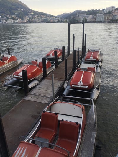 A fleet of bright red pedal boats are docked on a wooden pier extending into Lake Lugano, Switzerland. The boats, designed to resemble vintage cars, are lined up in the water. The city of Lugano is visible in the background, with buildings climbing up a lush green hillside. The sky is overcast with a soft, diffused light. One boat prominently displays the number "10" and the word "FORSA". Another boat has "2 FORSA" visible.