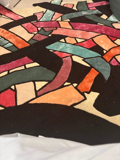 The image shows a close-up of a colorful textile with an abstract geometric design. The design features irregular shapes and lines in various colors, including shades of orange, pink, red, teal, green, and beige, outlined in black. The textile appears to be a rug or a decorative pillow. The setting is unknown but the mention of Arinj, Armenia is irrelevant to the content of the image itself.