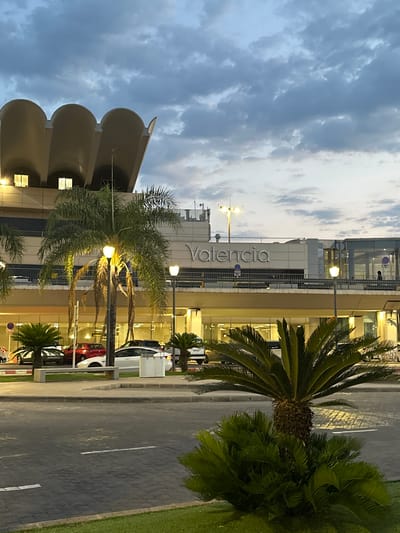 This image depicts the exterior of a modern building with the word "Valencia" prominently displayed in large, silver lettering. The building appears to be an airport terminal or a large commercial establishment, characterized by its glass facade and clean lines. Several palm trees and cycads are visible in the foreground and along the side of the building, suggesting a warm or Mediterranean climate. The ground is paved with dark stones and asphalt, with white road markings visible.
Several cars are parked along the side of the building, indicating that this is an accessible area. The lighting suggests it is either early morning or late evening, with artificial lights illuminating the building and its surroundings. The sky is cloudy, with a mix of light and dark gray clouds, hinting at potentially overcast weather. There are no discernible people in the image, and no specific activity or event is taking place.
The overall impression is one of a functional, modern architectural space set against a backdrop of greenery and urban infrastructure. The word "Valencia" is the only visible text. The scene is calm and static, without any indication of movement or immediate action. The time of day, combined with the artificial lighting and cloudy sky, creates a somewhat subdued atmosphere.