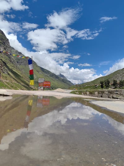 This is a low-angle, wide shot of a reflection of prayer flags and a sign in a puddle of water on a road in Koksar, India. The prayer flags are multicolored and hang on a pole. The sign is red and white and has text on it. The puddle reflects the flags, the sign, and the sky, which is blue with white clouds. The road is made of asphalt and gravel. In the background, there are mountains with green slopes. The scene appears to be a mountainous area in India.