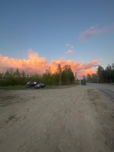 The image, correctly oriented, depicts a serene outdoor scene during either sunset or sunrise, characterized by a strikingly colorful sky. The upper left portion of the sky is clear blue, gradually transitioning to a vibrant display of orange, pink, and yellow clouds that stretch across the horizon on the right, bathed in the warm, soft light of the golden hour.

In the foreground, a wide, unpaved, dusty ground occupies the left side of the frame, interspersed with sparse patches of green vegetation. Two vehicles are parked on this ground: a dark-colored car and a maroon van, possibly a Mercedes-Benz Sprinter.

A dense line of trees, including both coniferous and deciduous types like birch, forms a prominent horizontal band across the middle ground, some appearing silhouetted against the bright sky. A paved road runs alongside the unpaved area, bordering the treeline. Near the road and trees, a dark green utility box or small shed is visible, with a streetlight pole extending upwards above the treeline.

No people are visible, and no specific activity is taking place beyond the presence of the parked vehicles. The scene conveys a tranquil, semi-rural or rural setting, consistent with the provided location context of Высокий, Russia. There is no visible text in the image.