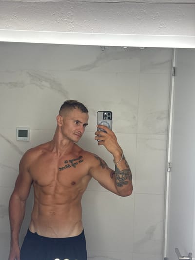 A man with short, spiked blonde hair and a muscular physique takes a mirror selfie in a brightly lit bathroom. He is shirtless, revealing tattoos on his chest and left arm. He is wearing dark athletic shorts. The bathroom walls are tiled with a white marble pattern, and a digital display is visible on the wall behind him. The man is holding a smartphone in his right hand, which is angled to capture the reflection. The scene appears to be in a modern hotel bathroom.
