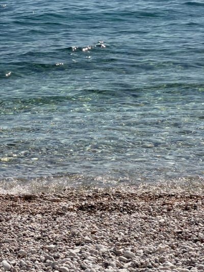 The image displays a serene aerial or overhead view of a clear, calm body of water meeting a pebble beach. The scene is dominated by the natural textures of the water and the shore.
The left side of the image features transparent water, revealing the seabed beneath in various shades of blue and green. The water surface is covered in gentle ripples, creating elongated patterns and reflections of sunlight that shimmer brightly, particularly closer to the shore. The clarity of the water suggests a shallow, undisturbed environment.
To the right, a pristine pebble beach stretches, composed of numerous smooth, rounded stones varying in size from small grains to larger, thumb-sized rocks. The pebbles exhibit a natural palette of grays, whites, and light brown hues. The demarcation between the water and the beach is distinct, with small splashes of foamy water visible along the immediate edge of the shore, likely from gentle waves.
The strong, direct sunlight indicates that it is daytime, possibly morning or early afternoon, contributing to the bright reflections on the water and the well-lit appearance of the pebbles. There are no people, animals, or man-made objects visible, emphasizing the natural beauty and tranquility of the scene. No text is visible in the image. This idyllic coastal setting is located in Grad Rijeka, Croatia.