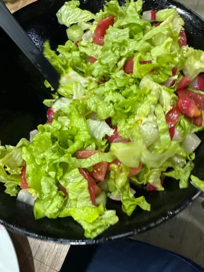 A close-up shot depicts a black bowl filled with a salad. The salad consists of lettuce, tomatoes, and radishes. A black serving utensil is partially visible at the bottom left of the frame. The salad sits on a wooden surface. A portion of a blue garment is visible in the upper right corner. The bottom right of the frame shows a white plate. The location is not discernible from the image.