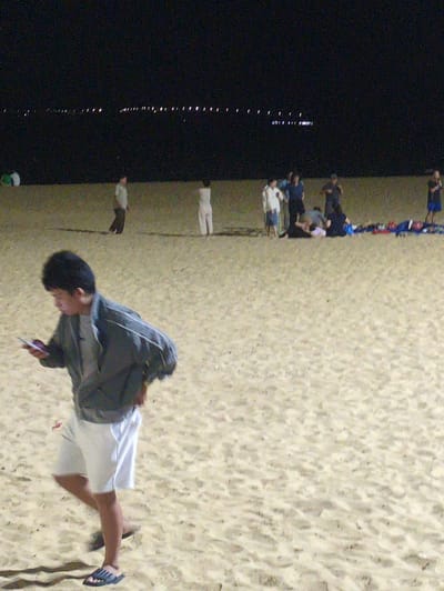 The image shows a beach at night in Quy Nhon Ward, Vietnam. A group of people is gathered on the sand near the water's edge. There are lights visible across the water, suggesting a cityscape in the distance. In the foreground, a person wearing shorts, sandals and a jacket is looking down at a phone while walking on the beach.