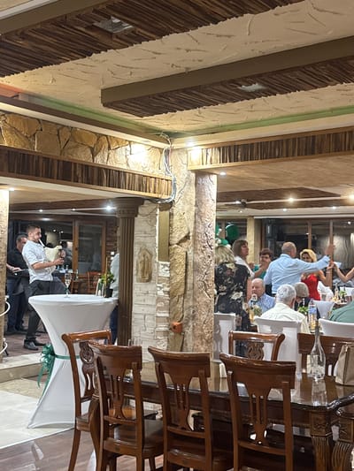 The image captures a lively indoor gathering, possibly a celebration or reception, set in a restaurant with rustic decor in Sredets, Bulgaria. The room features exposed stone walls, wooden beams on the ceiling with an unusual textured finish, and recessed lighting. In the foreground, a white cocktail table draped with a green ribbon is visible. To the left, a man in a light-colored shirt is playing a bagpipe-like instrument. A group of people are seen mingling and dancing in the background, suggesting a festive atmosphere. The tables are set with chairs, and bottles of water and glasses are present, indicating a meal or drinks service. The overall impression is one of a social event taking place in a well-decorated, traditional-feeling venue.