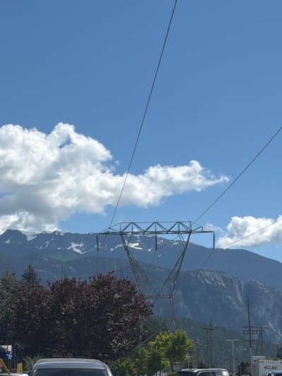 A high-angle, eye-level shot captures a mountainous landscape on a clear, sunny day. The sky is a vibrant blue, dotted with fluffy white clouds. In the foreground, a large metal power line tower stands prominently, with thick cables stretching across the frame. Beyond the tower, a range of majestic mountains rises, their peaks capped with snow. Lush green trees and a dark red-leafed bush are visible at the base of the mountains. In the lower part of the image, a silver car is parked, with other vehicles and utility poles suggesting a roadside or parking area. The scene appears to be located in Area D (Elaho/Garibaldi), Canada, based on the information provided.