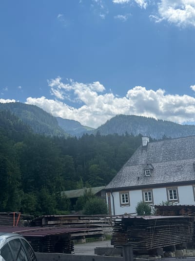 This image depicts a scenic view in Schönau am Königssee, Germany, featuring a traditional building with a grey, shingled roof and white-trimmed windows. The building is nestled against a backdrop of lush green mountains covered in dense forest. In the foreground, stacks of lumber are visible, suggesting a woodworking or construction-related area. The sky is a bright blue with scattered white clouds. The overall scene conveys a sense of rustic tranquility and natural beauty.