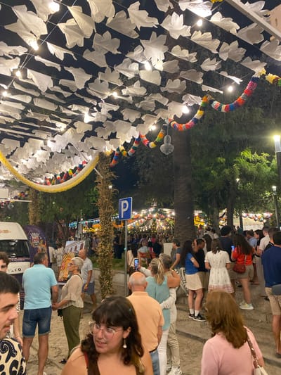 The image depicts a nighttime street scene in Tavira, Portugal, likely during a festival or local celebration. Above, strings of white, leaf-shaped decorations and colorful paper garlands are strung across the street, illuminated by overhead lights, creating a festive atmosphere. Trees and vegetation are visible in the background, with more decorative lights and banners further down the street. A parking sign with a blue "P" is mounted on a post on the right.

In the foreground and mid-ground, numerous people are gathered, milling about and interacting. A white van with "IVECO" and "TAVIRA" branding is parked on the left. The people are dressed in casual attire. Some individuals are standing in groups, others are walking, and some appear to be observing their surroundings or engaging in conversation. One woman in the foreground, wearing glasses and a white top, is looking towards the viewer. A couple in the mid-ground, a man in a blue polo shirt and denim shorts and a woman in a patterned top and green pants, are holding hands and looking at a display board. Another man in an orange shirt is also visible. The ground appears to be paved with cobblestones. The overall impression is one of a lively public gathering in an urban setting.