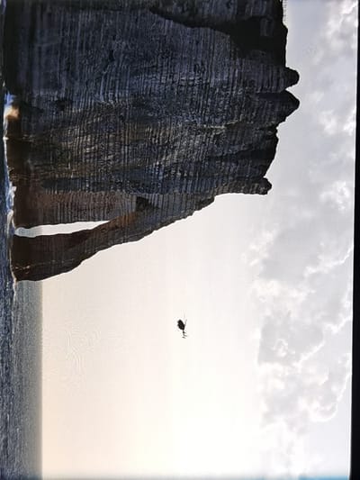 A helicopter is seen flying in the sky above the water, with a large rock formation behind it. The rock formation has a natural archway through it. The scene is likely Étretat, France, famous for its chalk cliffs. The image is taken from a low angle, looking up at the helicopter and cliffs. The sky is cloudy.