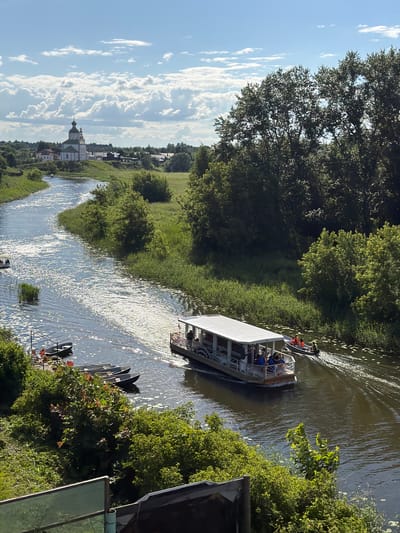This image captures a sunny day in Suzdal, Russia, with a wide, reflective river flowing through lush green banks. In the foreground, several small, empty boats are moored near a verdant embankment. Further up the river, a larger, white passenger boat with an open-air upper deck is cruising, carrying several passengers. A smaller dinghy with two individuals is following closely behind the passenger boat. In the distance, on a hill overlooking the river, a white church with a prominent green dome and bell tower stands as a focal point, surrounded by trees and other buildings, suggesting it is likely the Suzdal Kremlin or a similar historic religious complex. The sky is a vibrant blue, dotted with fluffy white clouds. The overall scene is one of serene summer activity in a picturesque Russian town.