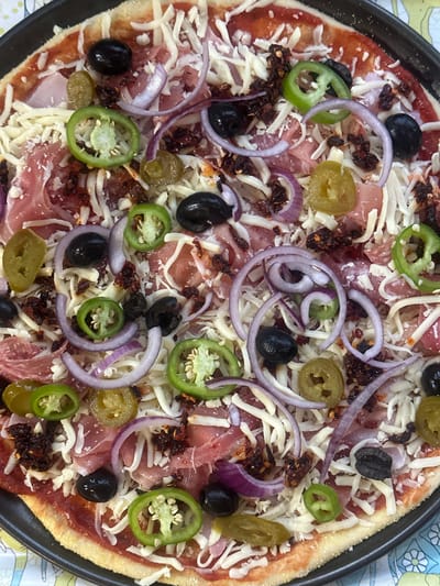 A close-up, high-angle shot depicts a pizza on a black pan. The pizza is topped with shredded cheese, red sauce, prosciutto, sliced green and yellow jalapenos, black olives, red onion slices, and small pieces of sun-dried tomatoes. The crust has a golden-brown hue. The pan sits atop a surface with a floral pattern. The image does not provide information about the location being in Wealden, United Kingdom or any people, actions, or interactions.
