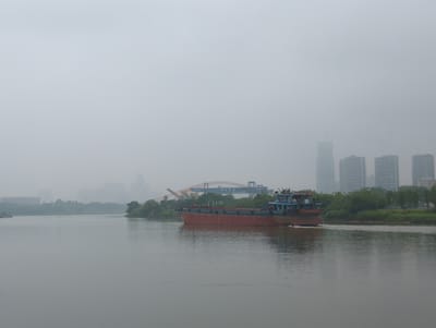 A cargo ship moves across a wide body of water. The ship is primarily red with a blue and gray upper structure. A faint wake trails behind the vessel.

The background includes an orange-colored bridge with blue gantry cranes. Further in the distance, buildings are visible, including high-rise structures. The location is Shunde District, China.

The overall atmosphere is hazy, obscuring details in the background. The water is relatively calm, reflecting the overcast sky. A tree-lined bank runs along the right side of the river.