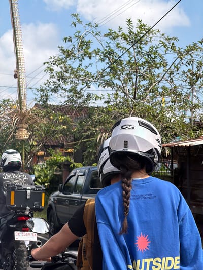 In Tampaksiring, Indonesia, on a bright, partly cloudy day, two individuals are riding on a motorcycle, viewed from behind. The motorcycle, a dual-sport model, is equipped with a black storage box on the rear. The rider in front wears a black t-shirt with a graphic and the text "Von Dutch" printed on it. The passenger behind has a long, braided brown ponytail and is wearing a blue sweatshirt with text that reads "DESIGNED FOR MOMENTS OF PAUSE" and a graphic with the word "OUTSIDE" below it. Both riders are wearing white helmets with black accents. The scene unfolds on a street lined with vegetation and buildings, suggesting a semi-urban or rural setting. In the background, a dark-colored pickup truck is visible, along with power lines and trees. To the left, a tall, ornate structure, possibly part of a cultural decoration, stands prominently. The overall impression is of a typical day on the road in Indonesia.