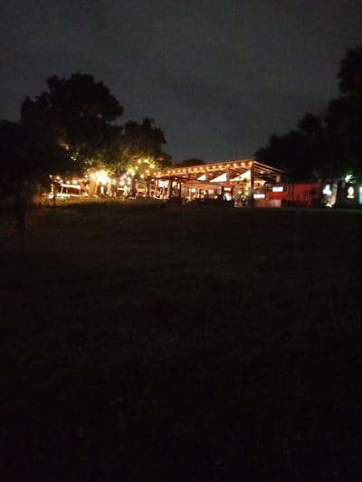This nighttime image depicts a lively outdoor setting, likely a restaurant, bar, or event space, in Maputo, Mozambique. The central feature is an open-sided structure with a wooden framework and a roof, brightly illuminated by numerous string lights strung across its beams. More string lights are draped among the dark silhouettes of trees that surround the building, creating a festive and inviting atmosphere.

Underneath and around the lit structure, several indistinct figures of people are visible, appearing to be engaged in social interaction, either seated or standing. To the right of the main structure, there is a reddish-orange container or smaller building, possibly a kiosk or food truck, also with some illumination.

The foreground of the image consists of a wide expanse of dark grass, almost entirely obscured by shadow, which starkly contrasts with the illuminated area in the mid-ground. The sky above is dark and overcast, characteristic of a cloudy night, further highlighting the artificial light sources. The scene conveys a sense of relaxed enjoyment and communal gathering. No visible text is discernible in the image.