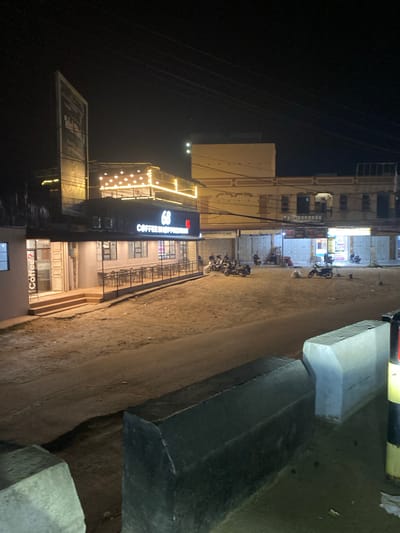 The image depicts an outdoor street scene at night in Seuneubok Teungoh, Indonesia, illuminated by artificial lights. The sky is dark, indicating the time of day.
On the left side of the frame, a row of multi-story buildings lines the street. A prominent building features a large, brightly lit sign that reads "COFFEE HOPPERIAN" in white and red lettering. Above this sign, an area is adorned with string lights, creating an inviting glow. Below "COFFEE HOPPERIAN," another sign or window displays "Coffee?". Further down the street, another building has a sign with the text "AL-BAROKAH," and partially visible below it is the word "CLINIC." Overhead, multiple utility lines are visible against the dark night sky.
The ground is an unpaved or dirt road that recedes into the distance. Several motorcycles are parked along this road, suggesting activity within the buildings. On the right side of the frame, concrete blocks or barriers line the edge of the road, and a yellow and black striped barrier is partially visible in the upper right.
The scene portrays a commercial or semi-commercial area during the evening, with businesses open and their signs lit. There is no specific activity or event captured, but the parked vehicles imply patronage of the establishments. The overall atmosphere is one of a quiet but active street at night, typical of an urban or semi-urban setting.