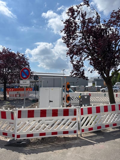The image captures a construction site in Mannheim, Germany, on a cloudy day. The foreground is dominated by temporary white and red construction barriers, casting shadows on the asphalt. Behind the barriers, a white modular structure with a dark door stands prominently. To the left, a circular road sign with a red border and a blue background displays two white arrows pointing in opposite directions, indicating no entry or exit except for authorized vehicles. Below this sign, a rectangular white sign reads "Ausser Werkstransport" (Outside of plant transport).
Further back, a large orange and white construction bin is visible, along with several large metal containers. Two trees with dark red foliage frame the scene on either side. In the background, a building with a grey facade and a green roof is partially visible, along with silver cars and a streetlight. The sky is blue with scattered white clouds, suggesting fair weather despite the overcast appearance. The overall impression is one of ongoing construction work, with clear signage indicating traffic restrictions.