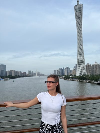 A full-body outdoor shot shows a young Caucasian woman with long brown hair tied back with black ribbons, wearing a white t-shirt and a black and white polka dot skirt. She is standing on a bridge, resting her left arm on a wooden railing. She is wearing black-rimmed glasses. In the background, a river flows through a city skyline featuring numerous skyscrapers and a distinctive, latticed white tower. The sky is overcast. The image appears to be taken in Guangzhou, Yuexiu District, China, given the prominent Canton Tower in the background.