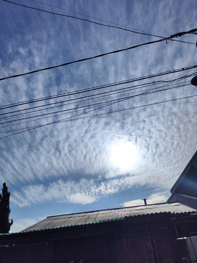 A daytime shot features a house roof below a sky filled with high, wispy clouds. The sun shines brightly, creating a halo effect amidst the cloud cover. Several power lines run across the top of the frame. The roof appears to be corrugated metal, and a tall, thin tree is partially visible on the left side. The house appears slightly obscured by shadows.