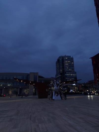 The image depicts an urban setting at dusk or night in Tiranë, Albania. The dominant subject is a tall, slender modern building on the left, illuminated with numerous lights from its windows, creating a distinct grid pattern against the dark sky. In the mid-ground, towards the right, stands a large, abstract sculpture composed of multiple reflective, angular metallic elements, resembling a fragmented starburst or crystalline form. This sculpture catches and reflects light from its surroundings.
The sky is a deep, uniform dark blue, indicating the evening hours. Below, a wide, flat paved area, likely a plaza or sidewalk, extends across the foreground and mid-ground. Other buildings with some lit windows are visible in the background and mid-ground, contributing to the city's nightscape. No specific activity or event is taking place; the scene is static, showcasing the urban environment after sunset. The overall mood is calm and illuminated by artificial lights. Faint text "udn" is visible on one of the buildings in the mid-ground.