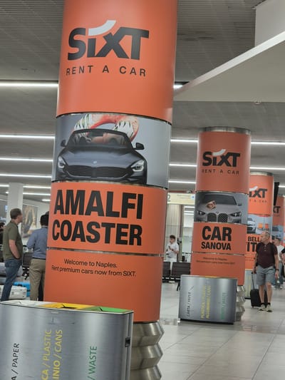 The image depicts an indoor scene, likely an airport terminal, with several large, orange cylindrical pillars displaying advertisements for SIXT Rent A Car. The pillars feature large SIXT logos and images of cars. One pillar prominently advertises "AMALFI COASTER" with a picture of a black convertible. Another pillar further back displays "CAR SANOVA". Smaller text on the pillars reads "Welcome to Naples. Rent premium cars now from SIXT." In the foreground, there is a multi-compartment recycling bin labeled in Italian: "CARTA / PAPER", "PLASTICA / PLASTIC ALLUMINIO / CANS", and "RIFIUTI / WASTE". Several individuals are visible in the background, including two men looking at the pillars, a young man holding a phone, and another man walking with a suitcase. The environment appears to be a public indoor space with a tiled floor and a suspended ceiling with lighting. The presence of the "Welcome to Naples" message on the SIXT advertisements suggests this location is in or near Naples, Italy, likely at the Naples International Airport (NAP).
