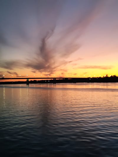The image depicts a sunset over a body of water, likely a river or bay, in the city of Shebunino, Russia. A bridge spans the water in the mid-ground, illuminated by a line of streetlights. The sky transitions from a pale blue at the top to vibrant oranges and yellows near the horizon, with wispy clouds catching the fading light. The water reflects the colors of the sky and the bridge lights. Silhouetted trees and buildings are visible along the far bank. The overall scene is serene and tranquil, characteristic of a twilight view.