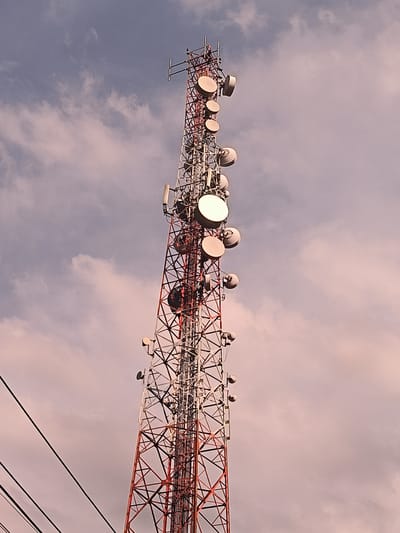 The image shows a tall telecommunications tower in Blangbitra, Indonesia. The tower is constructed of red and white lattice steel. It is equipped with numerous microwave dishes and other antennas of varying sizes and shapes attached at different heights. In the background, the sky is visible with light clouds. Power lines are visible in the lower left corner of the image.