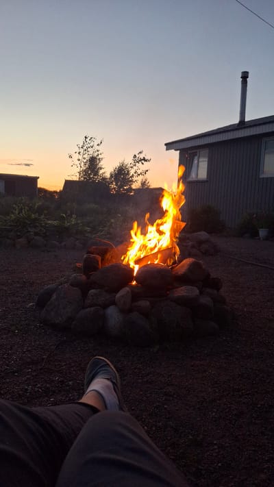In Kempelelevo, Russia, at dusk, a campfire crackles warmly within a ring of stones. The flames, vibrant orange and yellow, cast a flickering glow on the surrounding gravel and the dark, weathered siding of a nearby building. Silhouetted trees and vegetation are visible in the background against a sky transitioning from the pale orange of sunset to a deep twilight blue. In the foreground, a person's legs, clad in dark pants and wearing a shoe and sock, are casually positioned, suggesting they are enjoying the warmth of the fire. The scene evokes a sense of peaceful outdoor leisure.