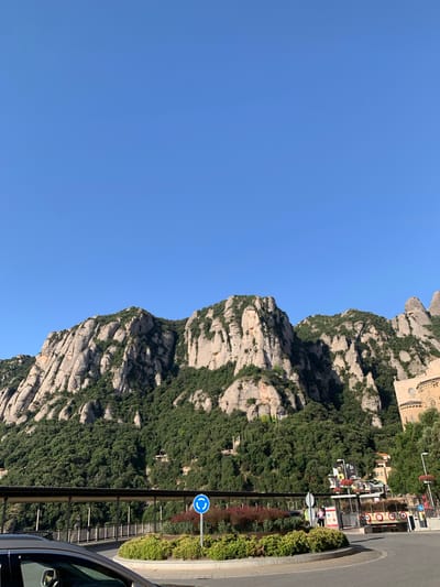 This image captures a view of the Montserrat mountains in Monistrol de Montserrat, Spain. The mountains are characterized by their unique, jagged rock formations covered in dense green foliage. In the foreground, a circular intersection is visible, featuring a roundabout with landscaping and a directional sign. A modern walkway or bridge runs horizontally across the mid-ground, with trees and buildings situated behind it. On the right side of the image, a stone building with a circular window, likely a church, is partially visible against the mountainside. The sky above is a clear, bright blue, indicating fair weather. A portion of a dark-colored vehicle is present in the lower left corner of the frame.