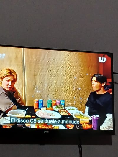 The image shows a television screen displaying a scene with two individuals seated at a table laden with food and drinks. On the left, a person with blonde hair is looking towards the camera, their right hand resting near their chin. On the right, a person with dark hair is looking away from the camera. The table features several bowls of food, plates of raw ingredients, and colorful cans, possibly beverages. A subtitle at the bottom of the screen reads "El disco C5 se duele a menudo". The background on the screen is a textured gold wall with some white speckles. The television is placed against a plain, dark gray wall. There is no visible indication of the city of El Espinal, Venezuela, within the image itself.