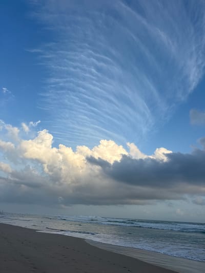 This is a landscape image of a beach with a dramatic sky. The scene is dominated by the sky, which takes up about two-thirds of the frame. On the left side, a vast expanse of clear blue sky is filled with wispy, streaky clouds that form a delicate, fan-like pattern. These clouds are thin and white, suggesting high altitude.
As the eye moves to the right, the sky becomes more dramatic with larger, denser clouds. A towering formation of cumulus clouds, with bright white tops and darker, shadowed undersides, juts into the frame from the right. These clouds appear to be illuminated from the side, possibly by the sun. Further to the right, the sky is overcast with dark, gray clouds, suggesting an approaching weather system or the lingering effects of one.
The bottom third of the image shows a beach and the ocean. The ocean is calm, with gentle waves rolling onto the shore. The water appears to be a muted gray-blue, reflecting the overcast sky. The beach is a strip of sand, light brown in color, with a few faint footprints visible. The horizon line is not straight, giving the image a slightly tilted appearance.
The time of day appears to be late afternoon or early evening, given the lighting on the clouds and the overall mood of the scene. The weather is mixed, with patches of clear sky and areas of heavy cloud cover. The location appears to be a tropical beach, with the presence of the ocean and sand. There are no people or man-made objects visible in the frame. The overall emotion evoked by the image is one of awe and natural beauty, with a sense of impending change suggested by the contrasting cloud formations.