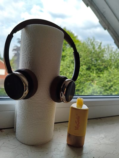 A roll of paper towels stands vertically on a windowsill. Headphones are placed over the top of the paper towel roll. To the right of the paper towel roll, a yellow disposable vape pen stands upright. The background shows trees and a cloudy sky through a window. The scene is indoors, likely in a residential setting.