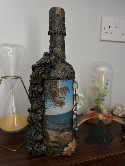 The image depicts an intricately decorated bottle on a shelf or table. The bottle is adorned with textured, dark green and brown material, possibly resembling sculpted clay or similar substance, covering most of its surface. A landscape image, featuring a beach with trees against a blue sky, is visible on the bottle. The bottle has a strand of beads wrapped around it.
Behind the bottle are various decorative objects. A glass dome encases some small plants. A figurine of a bird is visible, along with a small, black pyramid-shaped object. Below the bottle is an hourglass with a metal frame. A white electrical outlet is visible in the upper-left background.
There is no information in the image to suggest this object can be found in the city of Gorleston-on-Sea, United Kingdom.