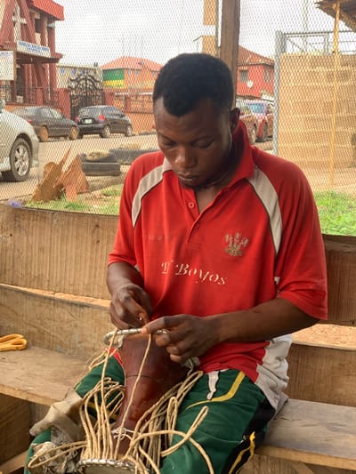 The image shows a man sitting on a wooden bench, wearing a red and white polo shirt with the word "Boys" printed on it, green and yellow pants, and a necklace. He is tying a string around a traditional Nigerian drum, which is made of wood and has a circular metal rim. The drum has a thick rope made of fibers wrapped around it. The image is taken in a street market setting in the city of Ondo, Nigeria (latitude: 7.249, longitude: 5.197). The man is focused on his work, his eyes fixed on the drum. In the background are cars, buildings, and a wire fence. There are several strands of fiber on the bench next to the drum. There are also yellow scissors lying on the bench in the lower left corner. The man's right hand is holding the string taut and he is using his left hand to tie it off. The image is taken at a slightly low angle, so the man's face is mostly in shadow.