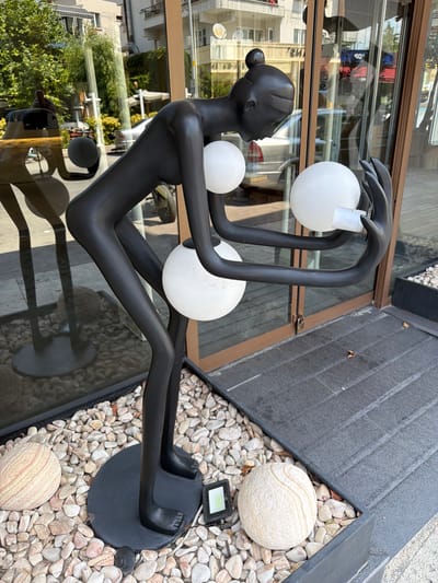 A black, abstract sculpture of a stylized human figure stands on a circular black base, surrounded by pebbles and decorative stones. The figure, which appears to be made of a matte material, is posed in a bent-over position, holding two large, white, spherical objects that resemble lamps or orbs. The left hand of the sculpture cradles one orb, while the right hand holds another, which is illuminated. The sculpture is positioned in front of a building with large glass doors and windows, reflecting trees, a car, and a motorcycle in the background. A small, dark-colored rectangular object, possibly a light fixture, is placed near the base of the sculpture. The ground in front of the sculpture is paved with dark, textured tiles. The overall scene suggests an outdoor display in a commercial or public space. The building in the background appears to be a shop or cafe, possibly located in Denizli, Turkey, given the architectural style and the presence of signage that may be in Turkish. The lighting indicates it is daytime, with sunlight casting shadows.