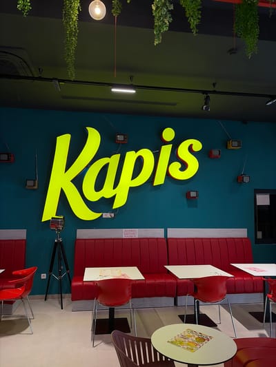 The image depicts the interior of a restaurant with a turquoise wall as the backdrop. A large, bright yellow neon sign with the word "Kapis" is prominently displayed on the wall. Several small, retro-style television sets with antennas are mounted on the wall, scattered around the neon sign. To the left of the sign, a vintage-style spotlight on a tripod is positioned.
The restaurant seating consists of red booth benches and red chairs. White tables are placed in front of the booths. In the foreground, a round table with a menu on it is visible. The floor is tiled in a light color. Hanging plants are suspended from the ceiling, adding a touch of greenery to the space. A light fixture with a round bulb is also visible in the upper part of the image. A sign with the text "SELF SERVICE" and "NARUCIVANJE I PLACANJE NA" is visible on the wall above the booth seating. The overall aesthetic suggests a modern and retro-themed dining establishment. The location is identified as Kovačko Polje, Montenegro.