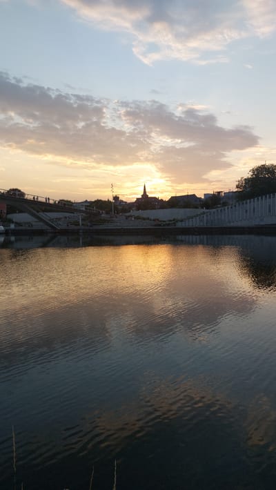 The image depicts a sunset over the River Sambre in Namur, Belgium. The sky is filled with a mix of pale blue and warm orange and pink clouds, with the sun partially visible behind a cluster of buildings on the horizon. In the foreground, the dark water of the river reflects the colors of the sky and the silhouette of the cityscape. A bridge with people walking across it spans the river in the mid-ground, and a tiered promenade lined with modern concrete structures is visible along the riverbank. The scene is peaceful, with the rippling water and the soft light creating a serene atmosphere.