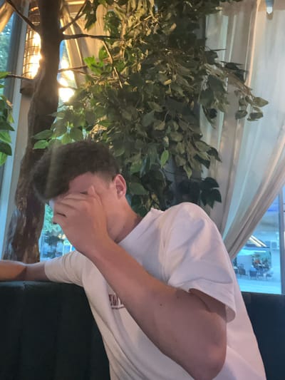 A young man with dark, curly hair is sitting on a dark green sofa, partially covering his face with his right hand. He is wearing a white t-shirt. The background features lush green foliage from what appears to be an artificial tree and a pale, sheer curtain. Behind the curtain, a brightly lit interior with tables and chairs is visible, suggesting the setting is a cafe or restaurant in the city of Gotse Delchev, Bulgaria. The lighting is warm and ambient.